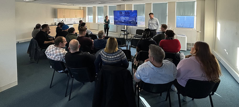 Team members at Boss Controls attend a company meeting in a bright office space. A presentation screen displays the Boss Controls logo and the text “Company Meeting December 2024,” while two speakers stand at the front addressing the group.
